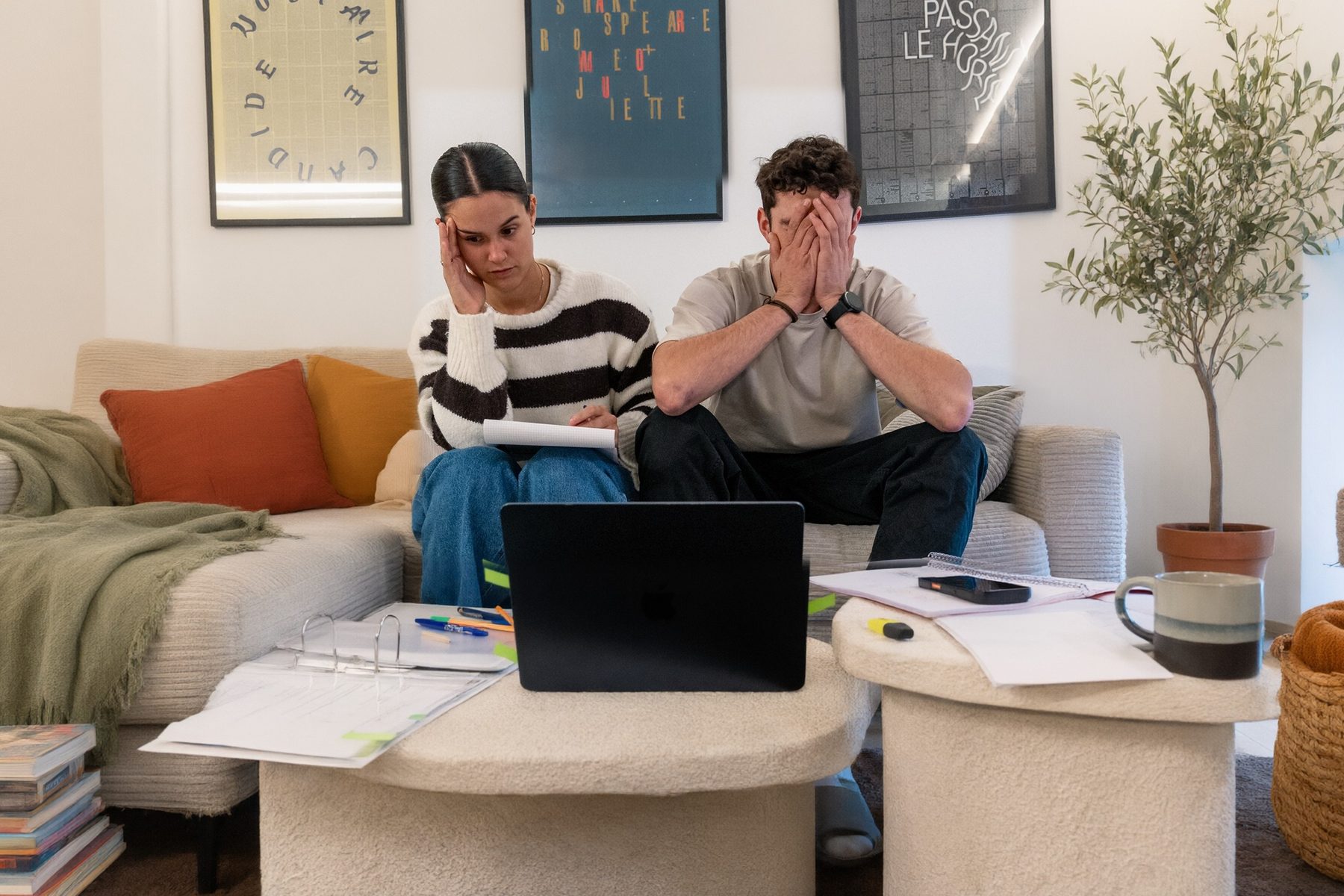 Un couple sur son canapé, la tête dans les mains, devant l'ordinateur, un classeur ouvert et des feuilles étalées sur la table basse